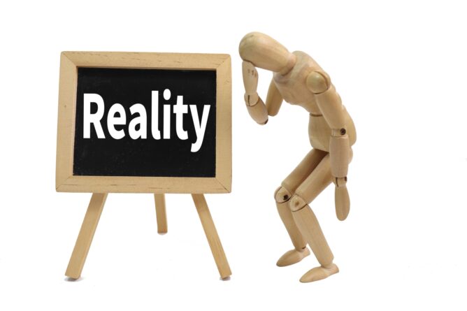 Wooden mannequin leaning toward a small chalkboard that displays the word Reality.