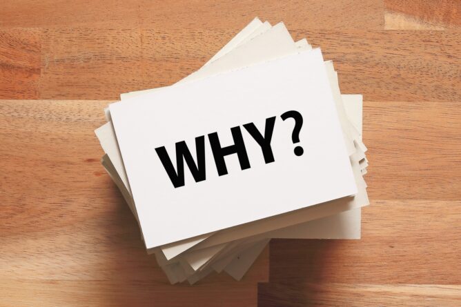 Stack of white cards on a wooden table with the word WHY? in bold black letters, suggesting a question or prompt.