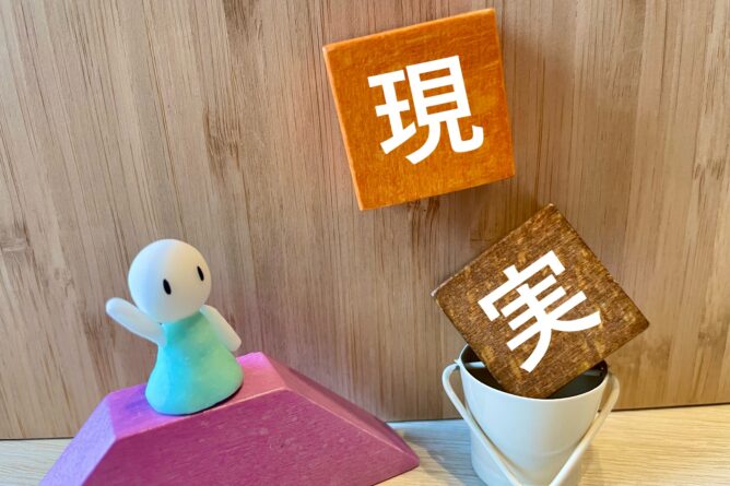 23671141_m | M Virtual(エムバーチャル) - Vライバーマネジメント事務所 Two wooden blocks with white Japanese kanji characters (現 and 美) rest near a small toy figure on a pink ramp against a wood backdrop.