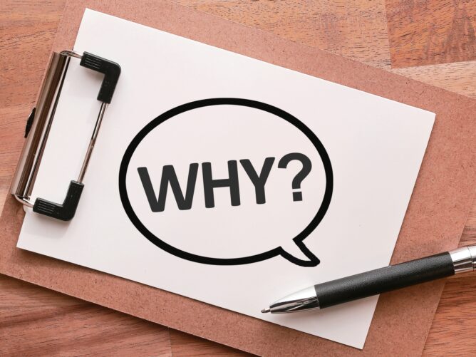 23777409_m | M Virtual(エムバーチャル) - Vライバーマネジメント事務所 Clipboard on a wooden desk with white paper showing a large WHY? inside a speech-bubble circle.