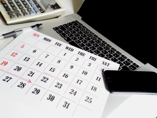 24526790_m | M Virtual(エムバーチャル) - Vライバーマネジメント事務所 Desk with a printed calendar showing a full month grid, a laptop nearby, a pen, and a smartphone resting on the right side.