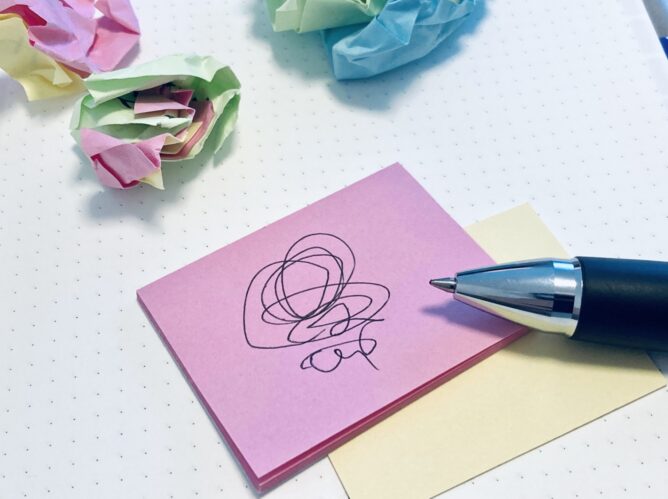 Pink sticky note with a black scribble, a blue pen nearby, and pastel notes on a white dotted desk with crumpled paper balls nearby.