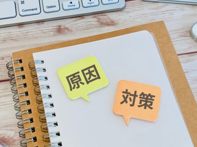 28564166_m | M Virtual(エムバーチャル) - Vライバーマネジメント事務所 Notebook with sticky notes showing the Japanese words for 'cause' (原因) and 'countermeasure' (対策) on a desk near a keyboard.