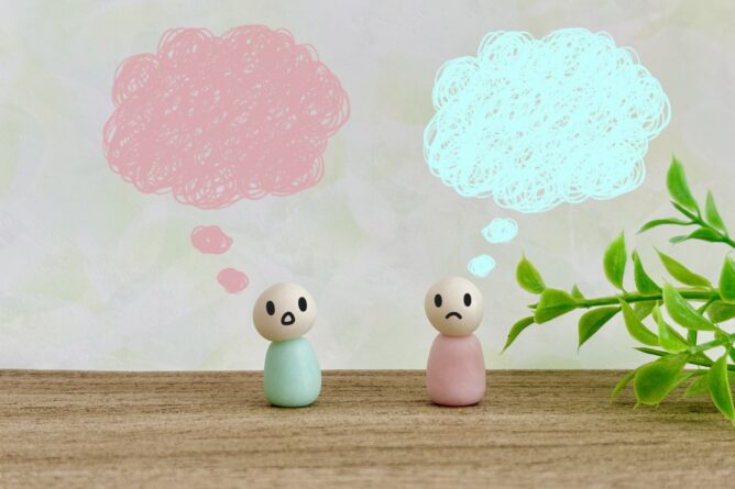 Two small pastel clay figures with sad faces sit on a wooden surface, each with a pink or blue thought bubble above them, a plant on the right.