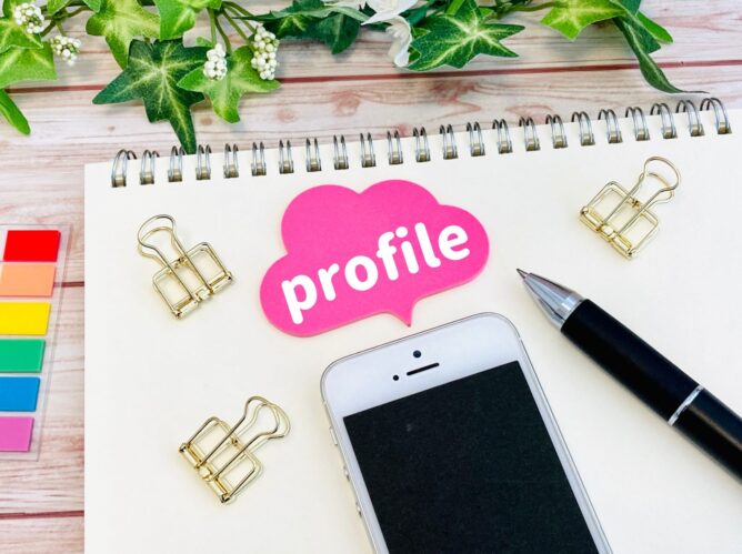 Flat lay of a desk with a pink'profile' cloud sticker, phone, pen, gold binder clips, color swatches, and green plants.