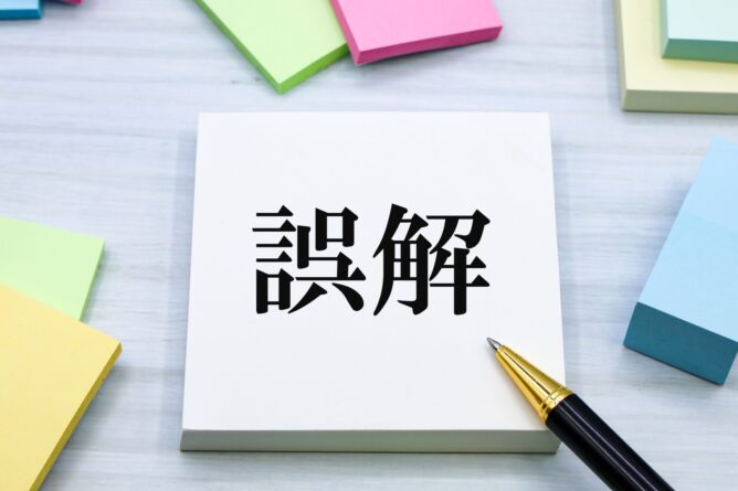 White notepad on a desk shows large black Japanese kanji'謎解' with colorful sticky notes and a gold-tipped pen nearby.