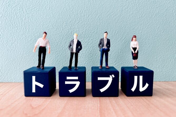 30149446_m | M Virtual(エムバーチャル) - Vライバーマネジメント事務所 Four miniature businesspeople stand on blue blocks; the blocks spell トラブル in white, meaning'trouble' in Japanese.