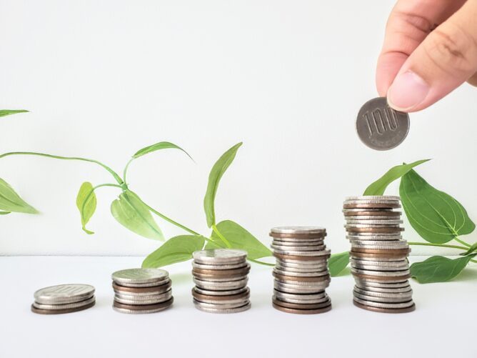 Coins are stacked from small to tall beside a green plant, illustrating growing savings or investment growth.