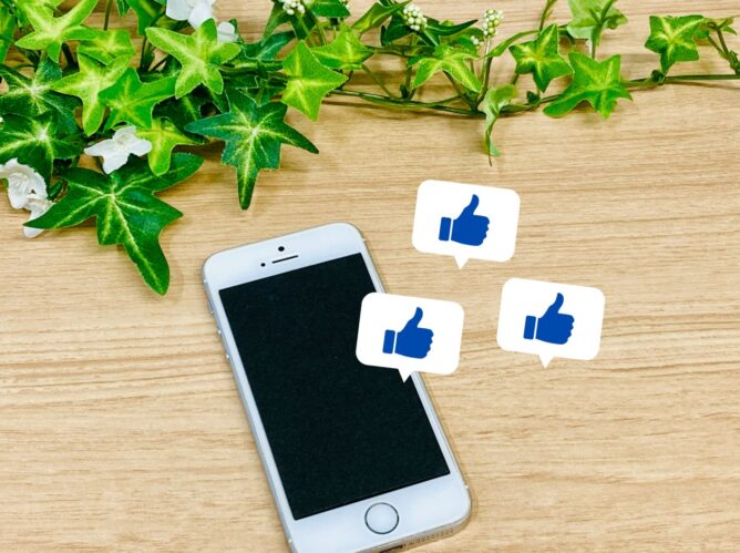 32132411_m | M Virtual(エムバーチャル) - Vライバーマネジメント事務所 Smartphone on a wooden desk with green ivy and white speech bubbles showing blue thumbs-up icons, representing social media likes.