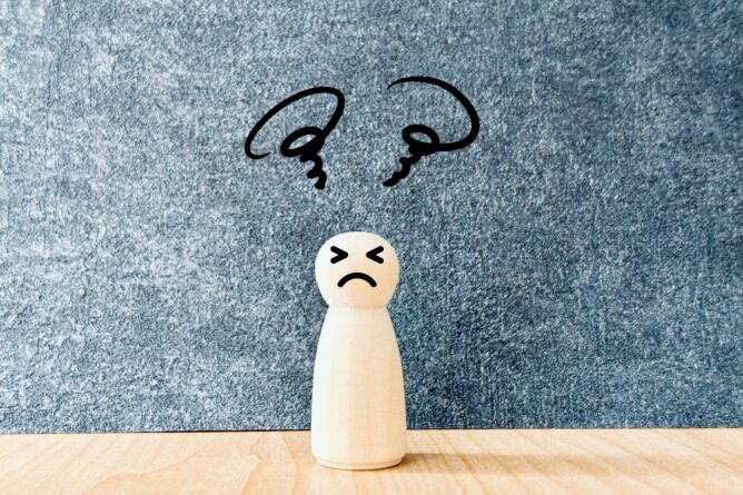 Small wooden figure with a sad expression on a light wooden surface, with squiggly black lines above suggesting worry or confusion against a blue textured background.
