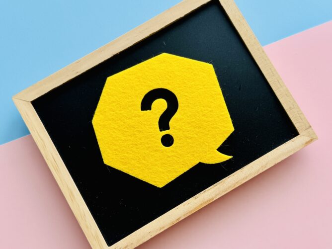 32731879_m | M Virtual(エムバーチャル) - Vライバーマネジメント事務所 Close-up of a yellow hexagon with a black question mark on a small chalkboard inside a wooden frame, set on pastel blue and pink backgrounds.
