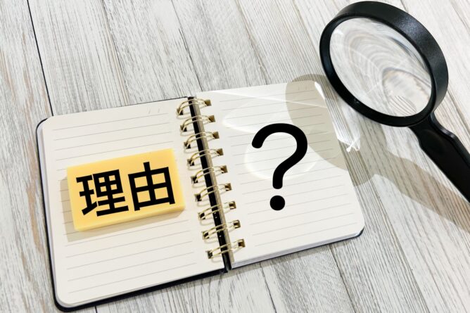 32947530_m | M Virtual(エムバーチャル) - Vライバーマネジメント事務所 Open spiral notebook with a sticky note bearing the Chinese word 理由 (reason) and a large question mark, plus a magnifying glass nearby on a wooden surface.