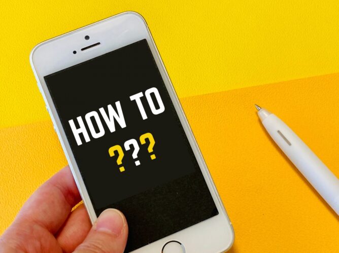 33107115_m | M Virtual(エムバーチャル) - Vライバーマネジメント事務所 Smartphone in hand with a black screen reading 'HOW TO ???' and yellow question marks on a bright yellow background.