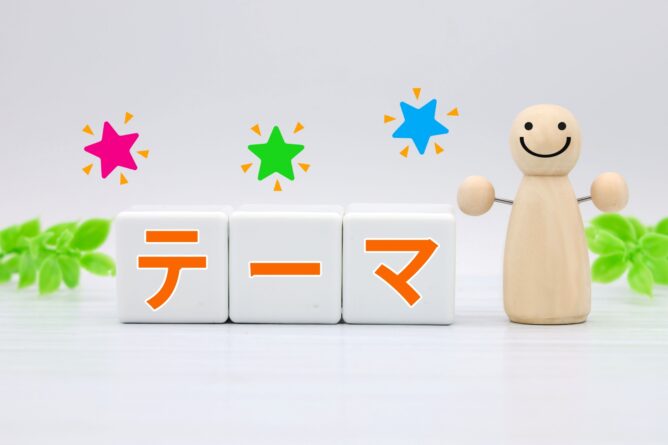 33120382_m | M Virtual(エムバーチャル) - Vライバーマネジメント事務所 Wooden smiling figure beside three white blocks spelling テーマ in orange, with pink, green, blue star bursts above on a pale background.