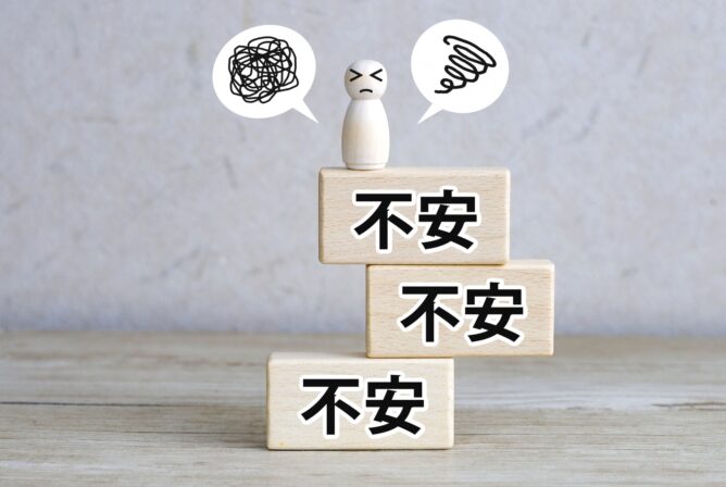 33253809_m | M Virtual(エムバーチャル) - Vライバーマネジメント事務所 Three wooden blocks stacked with the Chinese word 不安, a worried figure on top, and speech bubbles showing scribbles to depict anxiety.