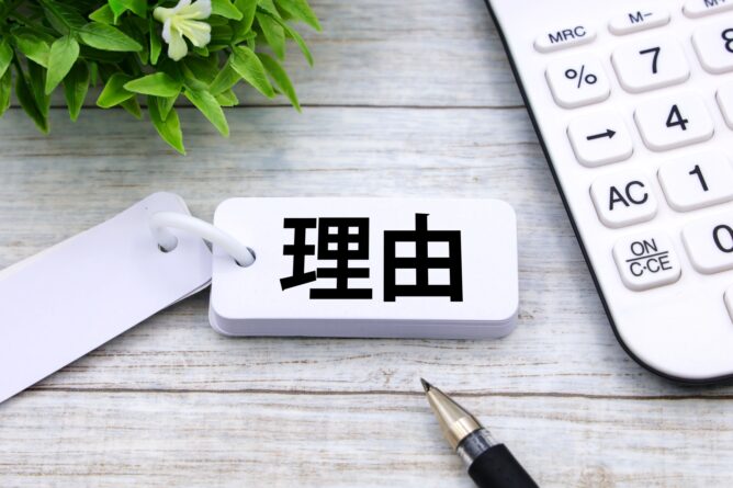 Tag with the Japanese word 理由 (reason) on a light wooden desk beside a calculator and a pen.