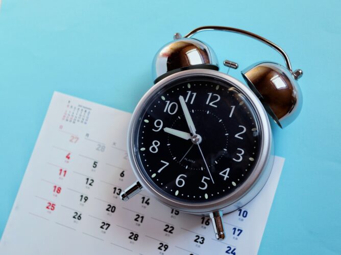 Black analog alarm clock with silver bells resting on a calendar page against a blue background.