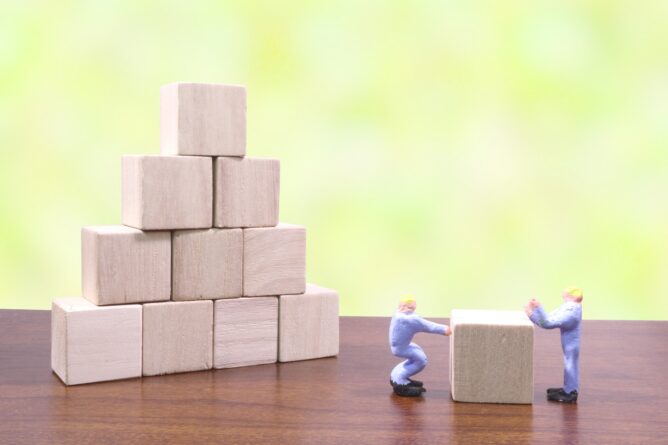 5142905_m | M Virtual(エムバーチャル) - Vライバーマネジメント事務所 Two miniature workers push a wooden block toward a pyramid of blocks on a table, showing teamwork in building.