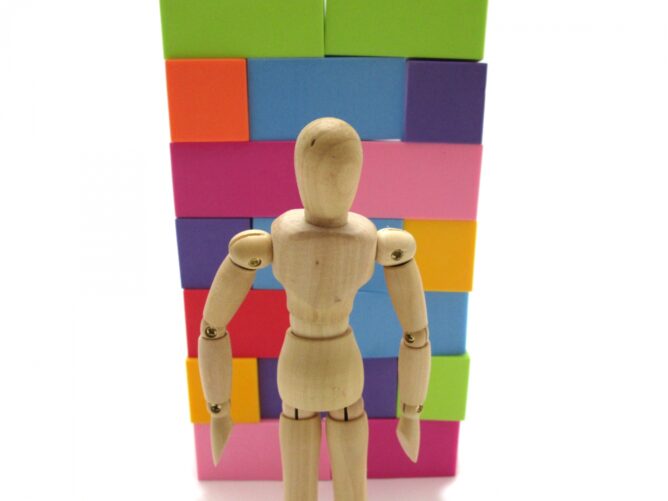 911079_m | M Virtual(エムバーチャル) - Vライバーマネジメント事務所 Wooden artist mannequin standing in front of a tall stack of colorful blocks in pink, blue, green, and purple hues.