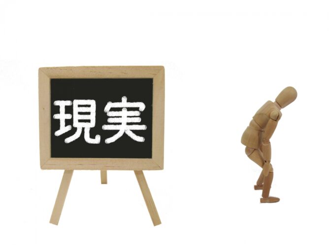 1950562_m | M Virtual(エムバーチャル) - Vライバーマネジメント事務所 Wooden mannequin gazing at a small chalkboard showing the kanji 現実 (reality).