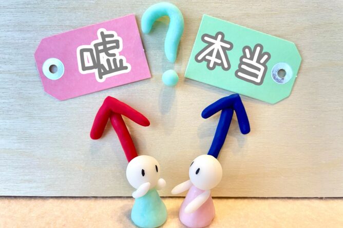 Two clay figures face each other under pink and green tags labeled 嘘 (lie) and 本当 (truth) with a question mark between them.