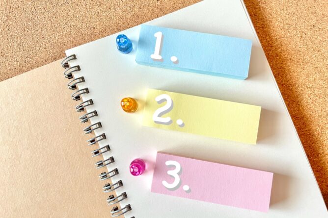 Three pastel sticky notes labeled 1, 2, 3 (blue, yellow, pink) placed on a spiral notebook against a corkboard background.