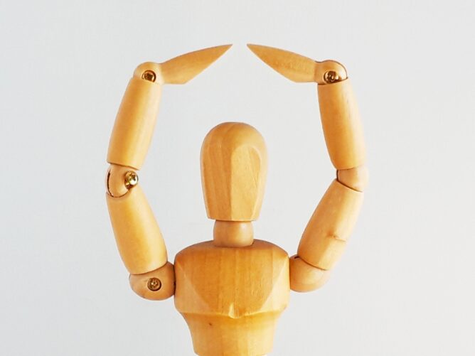 Wooden mannequin hand raising both fingers to form a heart shape against a light background