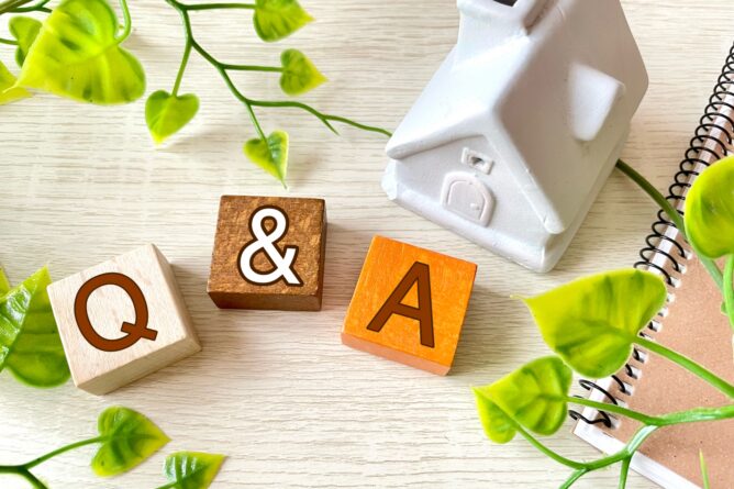 Wooden blocks spell 'Q & A' on a light desk, with a small white house model and green plants nearby.