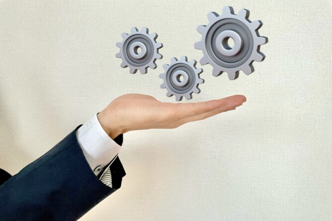 23349758_m | M Virtual(エムバーチャル) - Vライバーマネジメント事務所 Hand cupped under three floating gray gears, symbolizing automation and process optimization in the workplace.