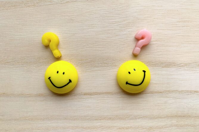 23695593_m | M Virtual(エムバーチャル) - Vライバーマネジメント事務所 Two yellow smiley faces with question marks above their heads on a wooden surface, symbolizing confusion or choice.