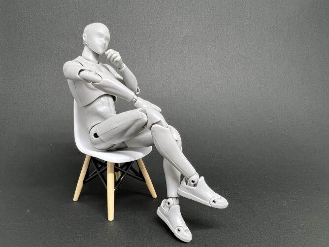 A gray humanoid mannequin sitting on a white chair with wooden legs, arms crossed and legs crossed, in a studio setting