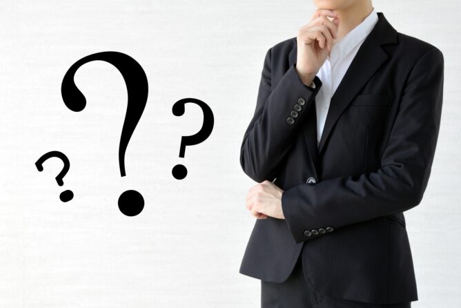 2596746_m | M Virtual(エムバーチャル) - Vライバーマネジメント事務所 Person in a dark suit with one hand on chin, large question marks floating to the left, conveying uncertainty or pondering a question.