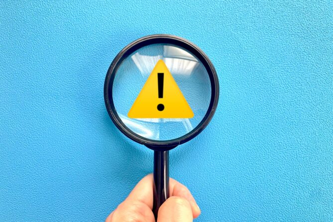 26026913_m | M Virtual(エムバーチャル) - Vライバーマネジメント事務所 Magnifying glass showing a yellow warning triangle with an exclamation mark against a blue textured background.