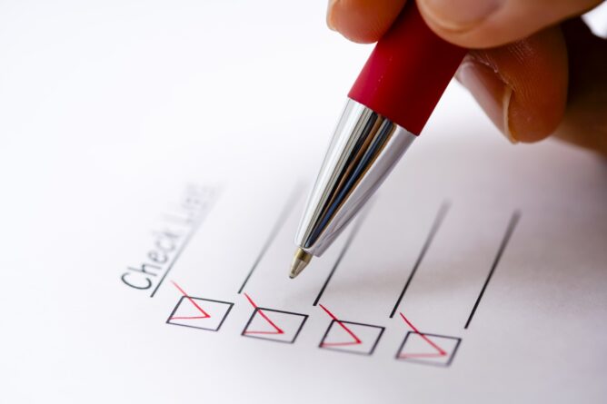 Close-up of a hand using a red pen to check off items on a printed checklist.