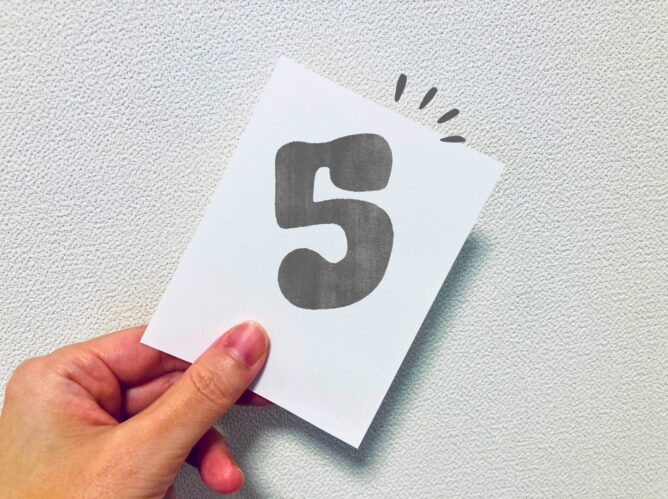 26787788_m | M Virtual(エムバーチャル) - Vライバーマネジメント事務所 Hand holding a white card with the bold number 5 on a light textured surface, angled at the corner.