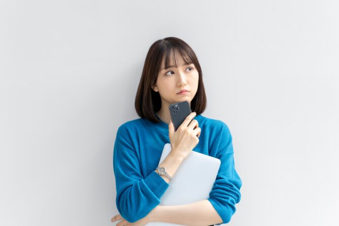 28151299_m | M Virtual(エムバーチャル) - Vライバーマネジメント事務所 Young woman in a blue sweater holds a smartphone to her chin while clutching a light laptop, looking thoughtful against a light wall.