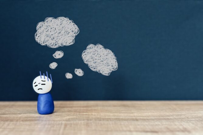 28639332_m | M Virtual(エムバーチャル) - Vライバーマネジメント事務所 Blue clay figure with a sad face sits on a wooden desk, with a blue chalkboard backdrop and white tangled thought bubbles above its head.