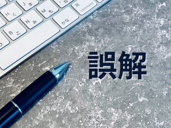 30585920_m | M Virtual(エムバーチャル) - Vライバーマネジメント事務所 Close-up of a white keyboard and blue fountain pen on a gray textured surface, with the Japanese word 誤解 ('misunderstanding') in bold to the right.