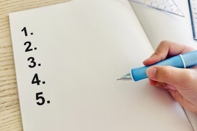 Hand holding a blue pen poised to write a five-item checklist on a blank notebook page.
