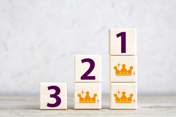 32115496_m | M Virtual(エムバーチャル) - Vライバーマネジメント事務所 Wooden blocks arranged in a staircase show numbers 3, 2, and 1, with orange crowns on the two right-side blocks, indicating a ranking.