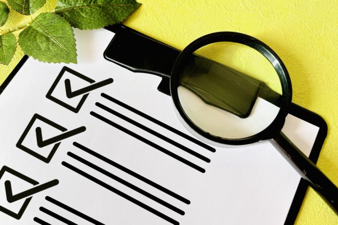 Checklist on a clipboard with checked boxes and a magnifying glass, green leaves in the corner, on a yellow background.