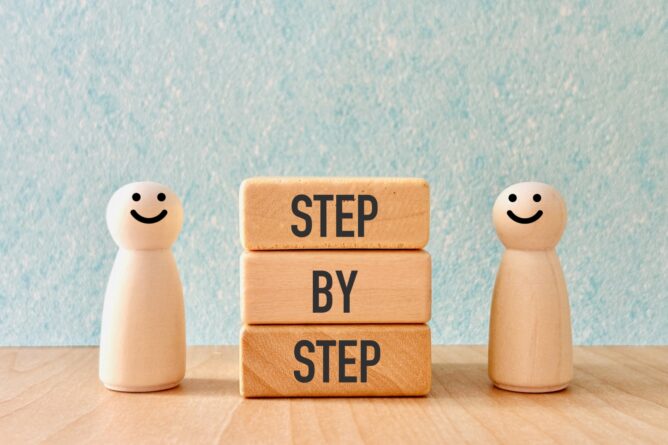 32916496_m | M Virtual(エムバーチャル) - Vライバーマネジメント事務所 Two smiling wooden figures stand beside three blocks that spell ‘STEP BY STEP’, conveying a step-by-step process concept.