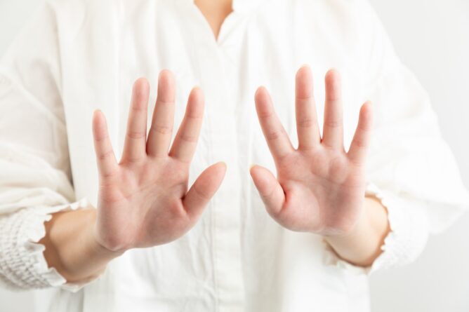 Person wearing a white shirt holds up both hands with palms facing the camera, signaling stop or attention.