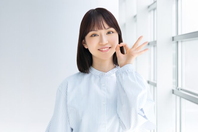 Smiling woman in a blue striped shirt making an OK sign with her hand by her face in a bright corridor with windows
