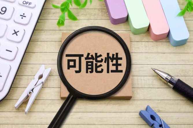 33651159_m | M Virtual(エムバーチャル) - Vライバーマネジメント事務所 Magnifying glass over a sticky note with the Chinese characters for 'possibility' on a wooden desk with office supplies nearby.