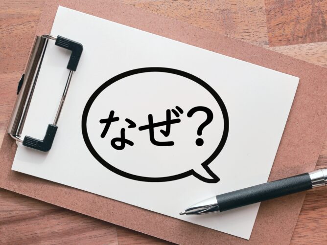 34296242_m | M Virtual(エムバーチャル) - Vライバーマネジメント事務所 Clipboard on a wooden desk with a white sheet featuring a large speech bubble containing the Japanese word なぜ? (Why).