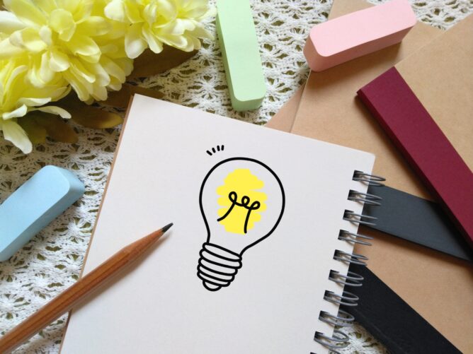 Notebook page with a yellow lightbulb sketch, surrounded by pastel erasers and pencils on a lace background.