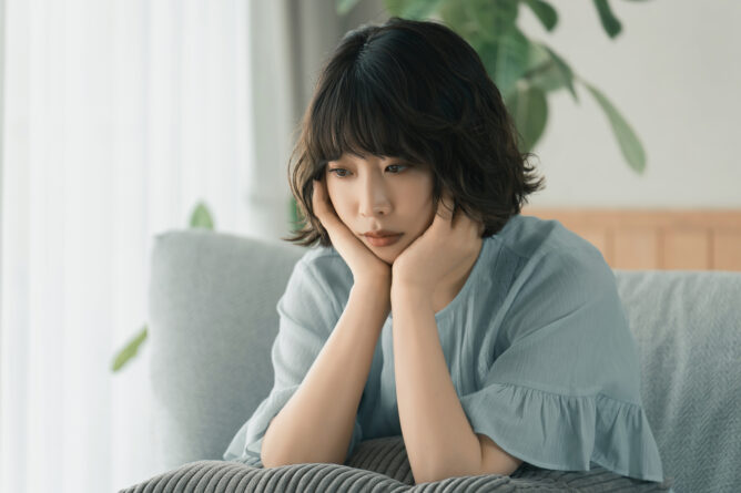 Woman sits on a gray sofa with hands cupping her face, looking downward in thought.
