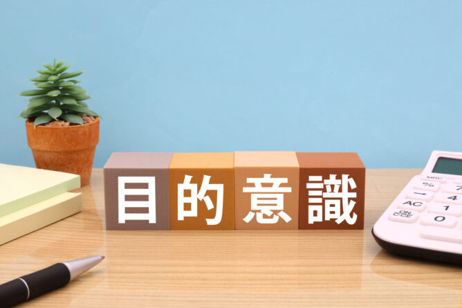 Desk scene with a potted succulent, four colored blocks displaying Chinese characters, a white calculator, and a pen on a wooden desk against a blue wall.