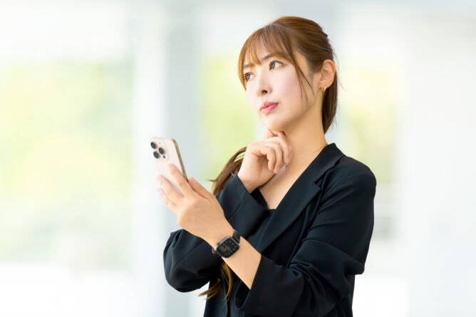 Professional woman in a black blazer holds a smartphone, looking thoughtful with her hand on her chin in a bright, blurred background office setting.